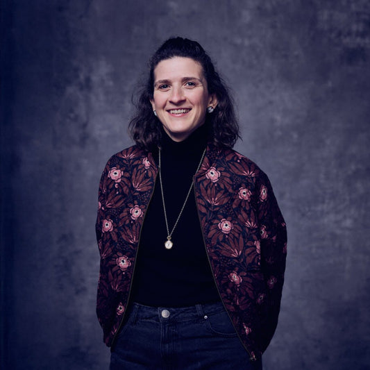 A professional photo of Jane Laverty, Communications Manager at Selvedge magazine. She is smiling, wearing a black t shirt with a patterned quilted jacket over the top, and is standing in front of a grey textured background.